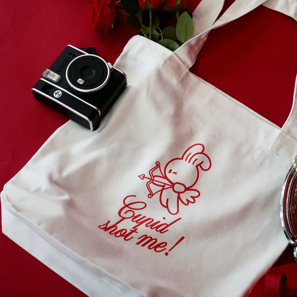 White tote bag with red text and bunny graphic, black camera on a red surface